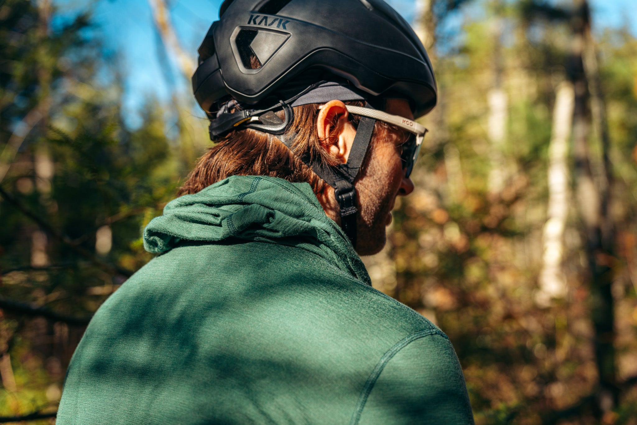 man standing in a forest, with a green hooded shirt and black helmet on.