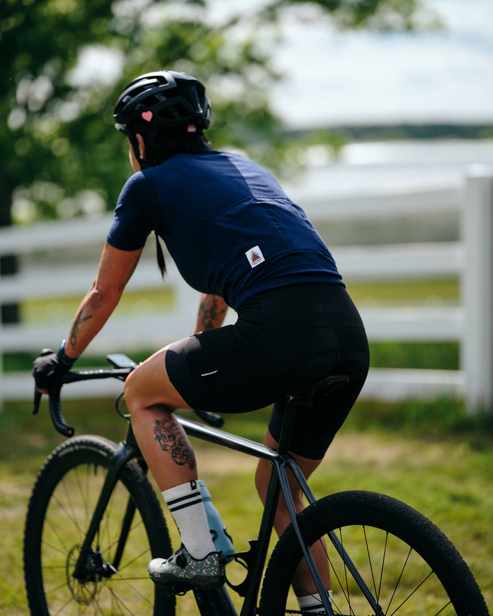 Pinebury Signature Merino Wool Sock in Natural. Female cyclist with tattoos riding away from the camera with a white horse fence in the background. She is wearing a blue short sleeve jersey and black bibs with white socks and colorful cycling shoes.