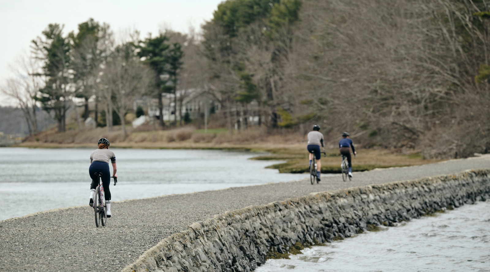 Three cyclists riding away from the camera on a gravel path between two bodies of water.