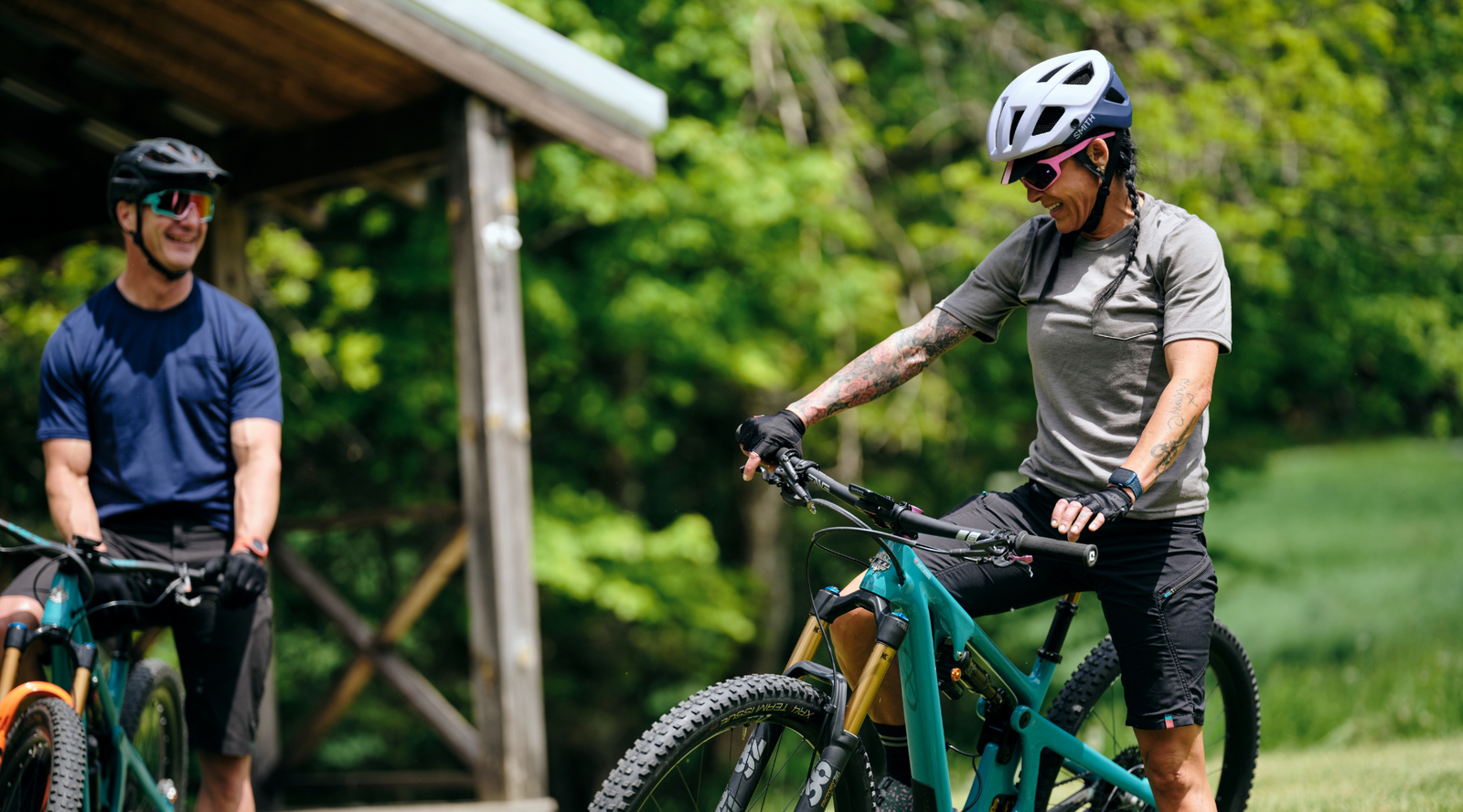 Pinebury Portland Merino Wool Tees in Atlantic Blue and Granite. Two mountain bikers one teal Yeti bikes, one male and one female, before a ride smiling in conversation.