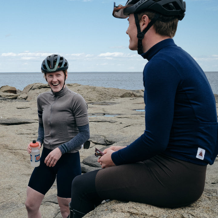 Pinebury Signature Merino Wool Arm Warmers in Grey. Male and a Female cyclist stopped on coastal rocks looking at each other with the female facing the camera. She is laughing while holding a water bottle and wearing a grey jersey with grey arm warmers.