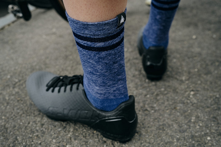 Pinebury Signature Merino Wool Sock in Blue. Close up rear view of cyclist wearing socks with black and grey lace up shoes, showing tag detail on top back of sock.