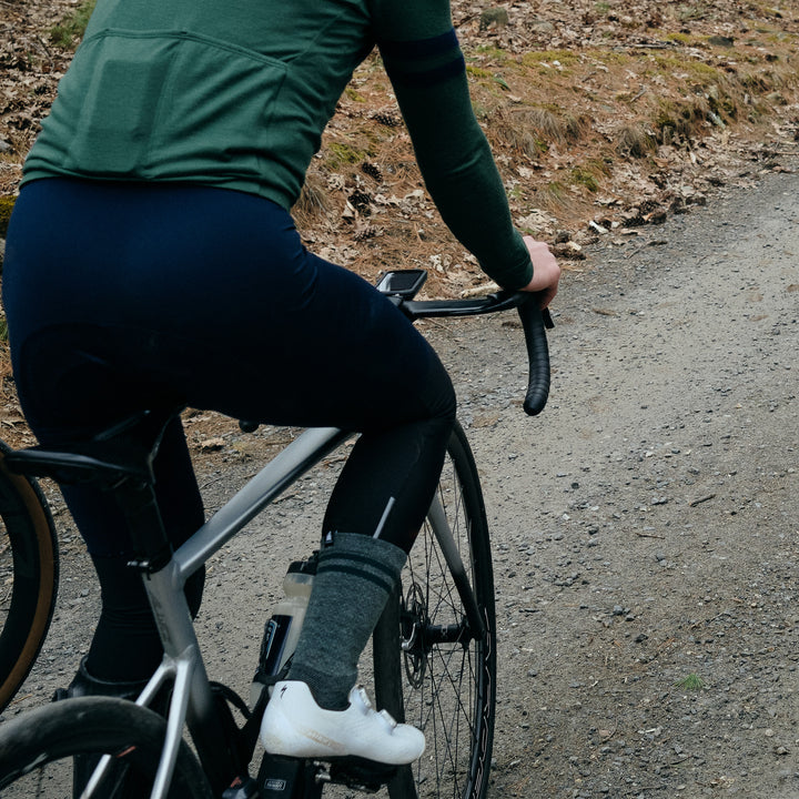 Pinebury Signature Merino Wool Sock in Loden Green. Cyclist riding up on a gravel road showing just their lower half, in green jersey, blue bibs and green socks with white shoes and a silver road bike. 