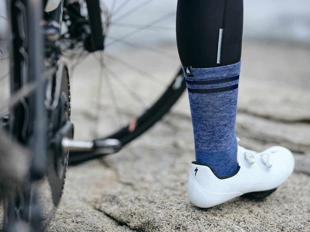 Pinebury Signature Merino Wool Sock in Blue. Rear view close up of cyclist standing on rocks with bike out of focus to the left. Wearing black leg warmers and white shoes, with a blue sock.