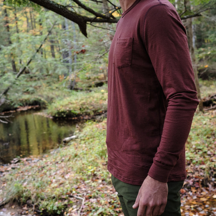 Pinebury Greenwood Long Sleeve Performance Tee in Brick Red, Photo of the upper body of a person standing in the woods looking at a creek in Maine