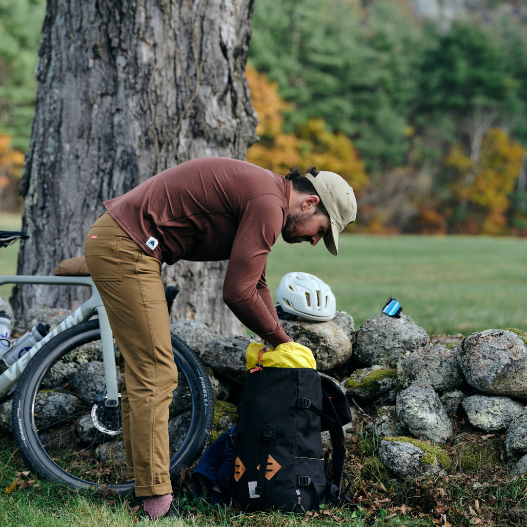Pinebury Greenwood Long Sleeve Performance Tee in Brick Red, Photo of the a man bending over pulling clothes out of a bag before a gravel ride