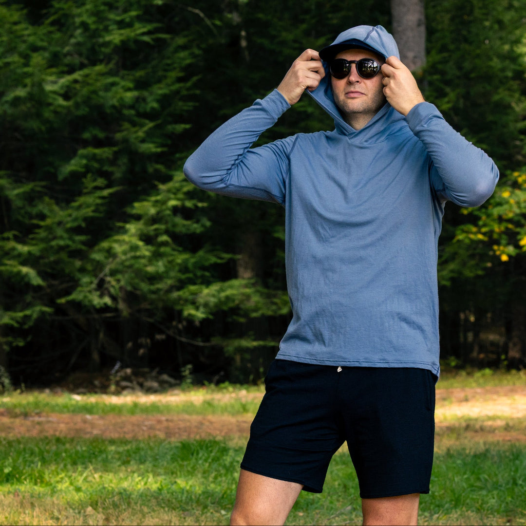 A man wearing a light blue long-sleeve hooded athletic top, black shorts, and sunglasses stands outdoors in a grassy clearing with a dense forest background, adjusting the hood of his shirt.