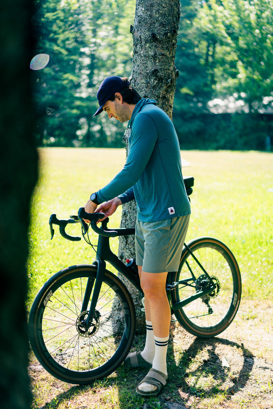 A man in a teal long-sleeve cycling jersey, gray shorts, and a dark cap stands next to a black road/gravel bicycle, leaning against a tree in a sunny grassy park.