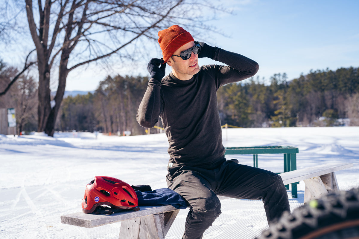 A man wearing an orange beanie, black sunglasses, a black long-sleeve shirt, and black pants sits on a snowy bench outdoors. A red bicycle helmet and blue jacket rest on the bench beside him, with trees and a snowy landscape in the background.