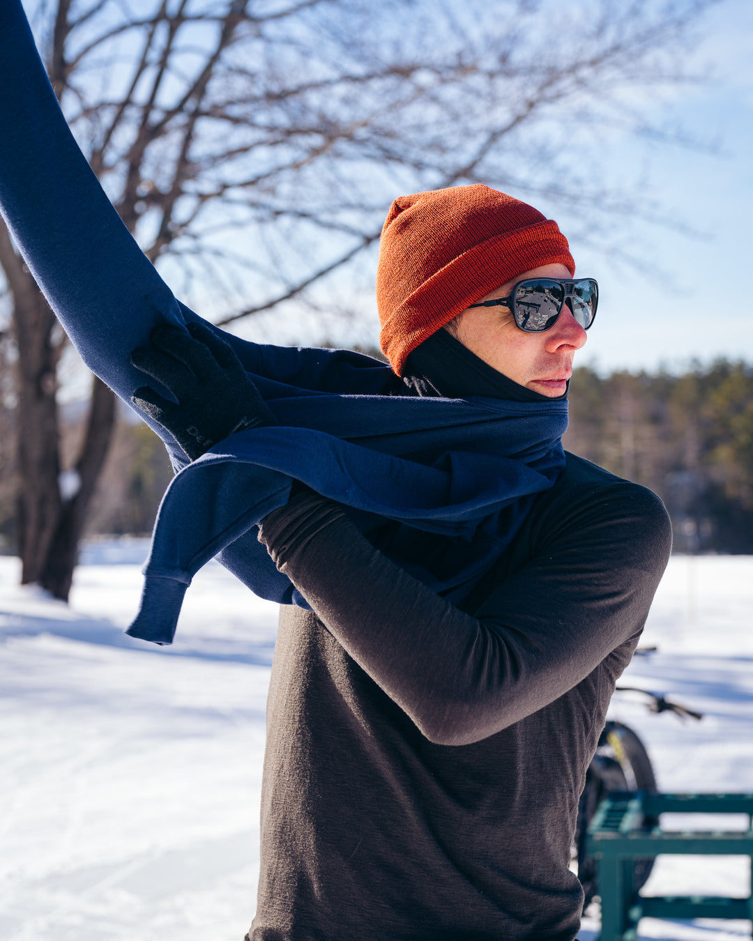 A person outdoors in a snowy setting wearing a dark navy long-sleeve top, an orange knit beanie, black sunglasses, and a black neck gaiter, while adjusting or wrapping a navy scarf or wrap around their neck.