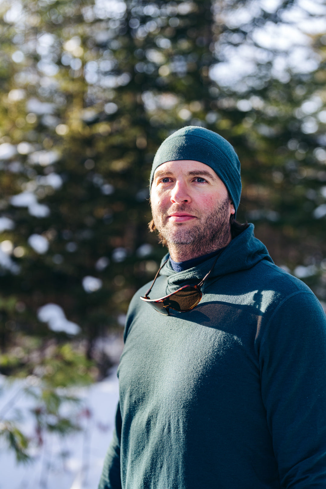 Man wearing a blue beanie and dark hoodie standing in a snowy forest