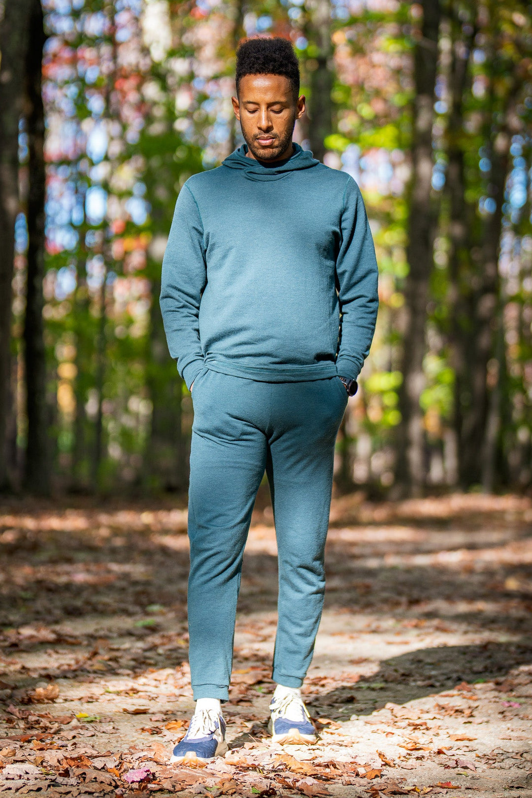 Person wearing a teal tracksuit standing in a forest with autumn leaves on the ground.