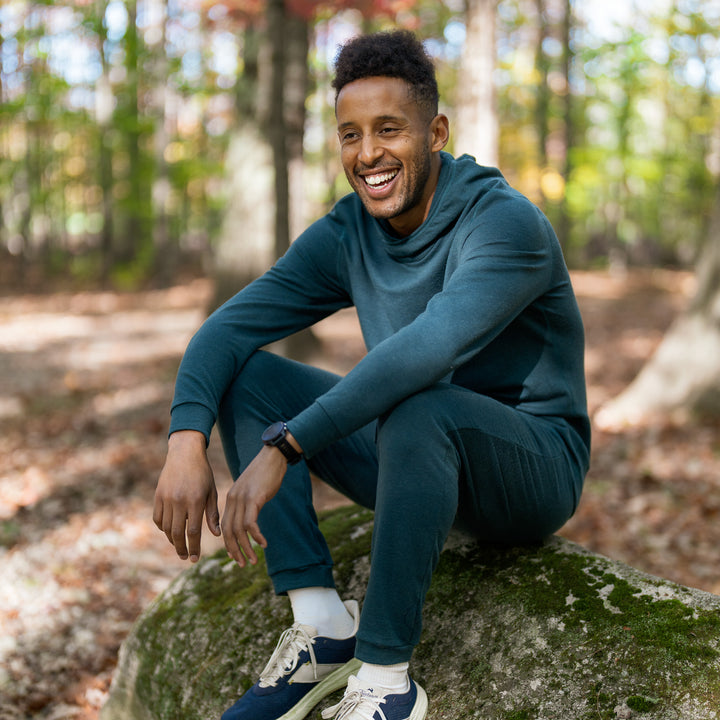 Man sitting on a rock in a forest wearing a teal tracksuit.