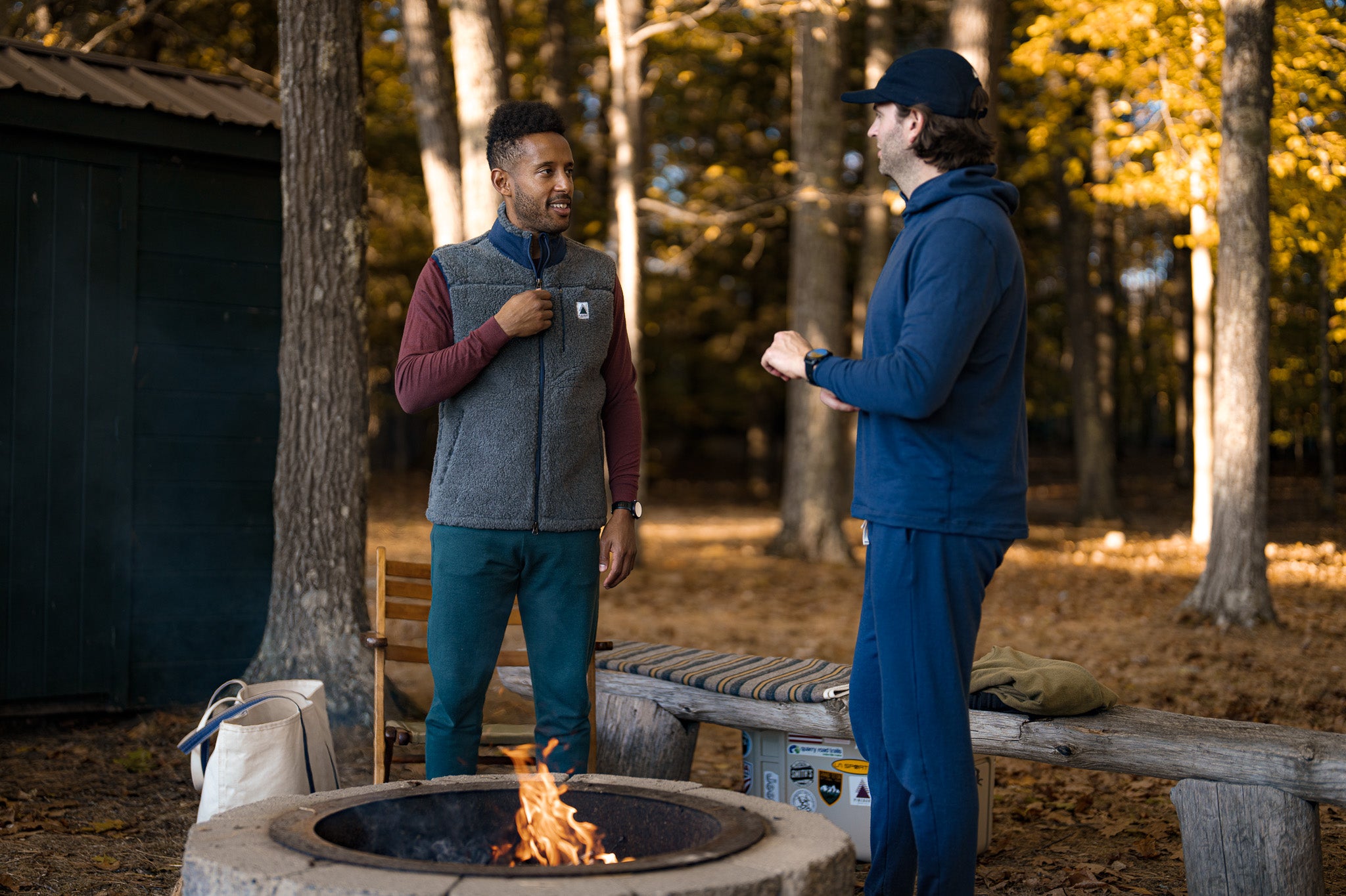 Two men standing around a fire pit in a forest setting
