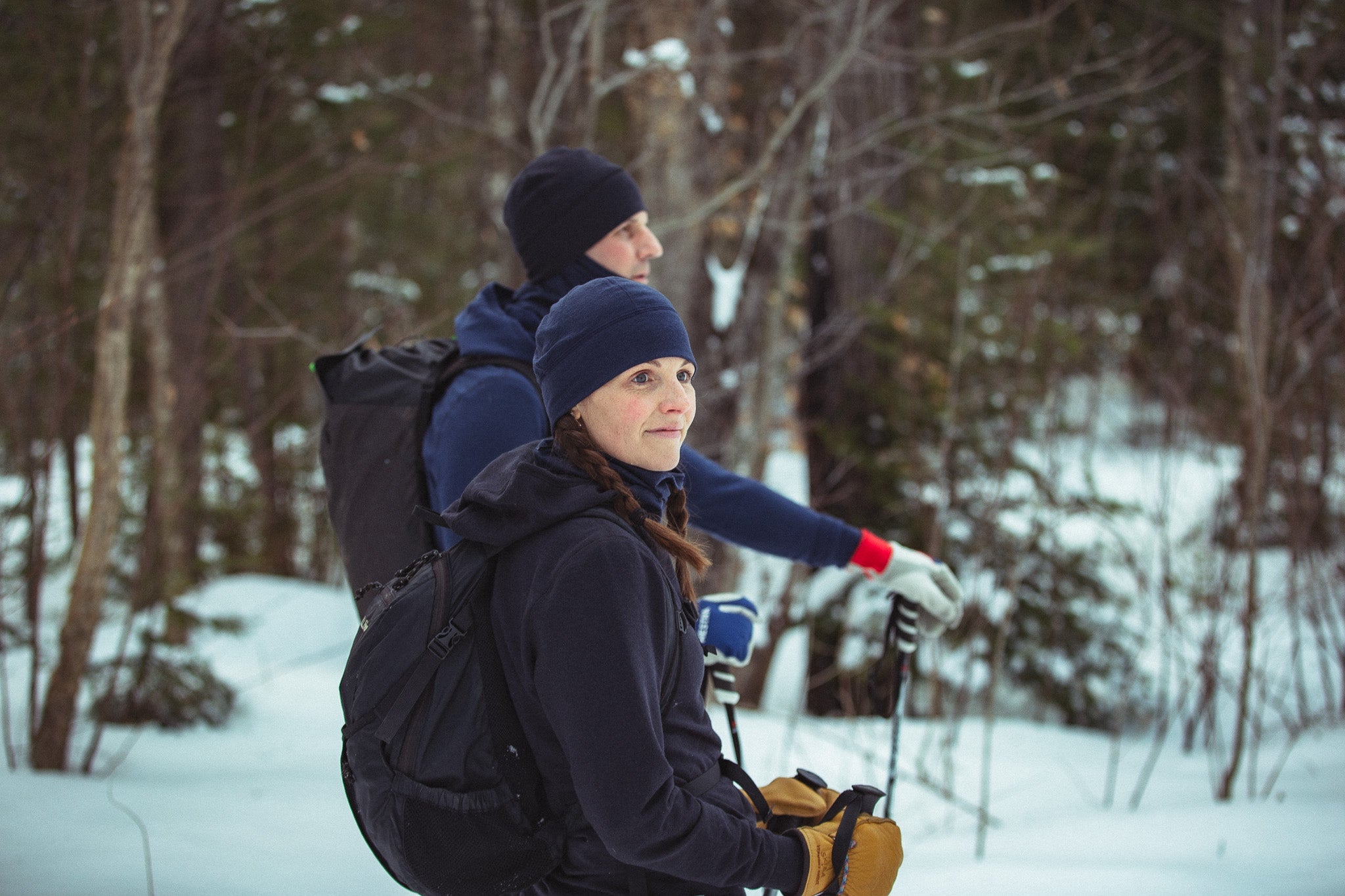 Two people hiking in a snowy forest with backpacks and ski poles.
