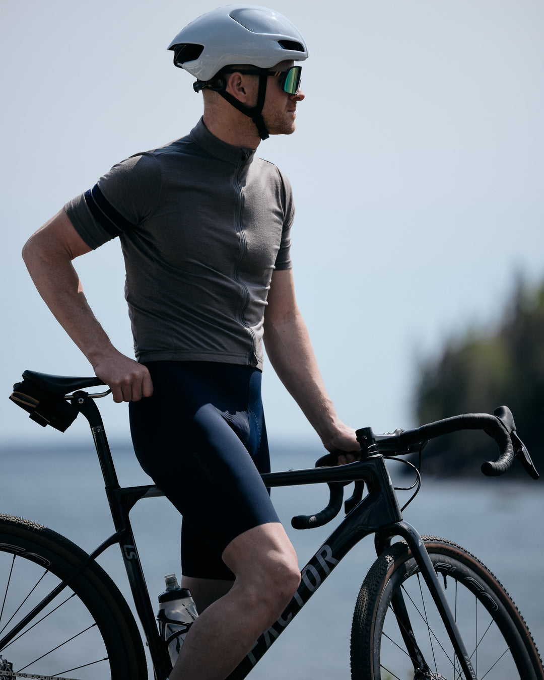 Pinebury Rangeley Long Sleeve Merino Wool Cycling Jersey in Granite. Male cyclist stopped on the coast standing over his bike looking to the side. Wearing a grey short sleeve jersey and blue bib shorts and white aero helmet.