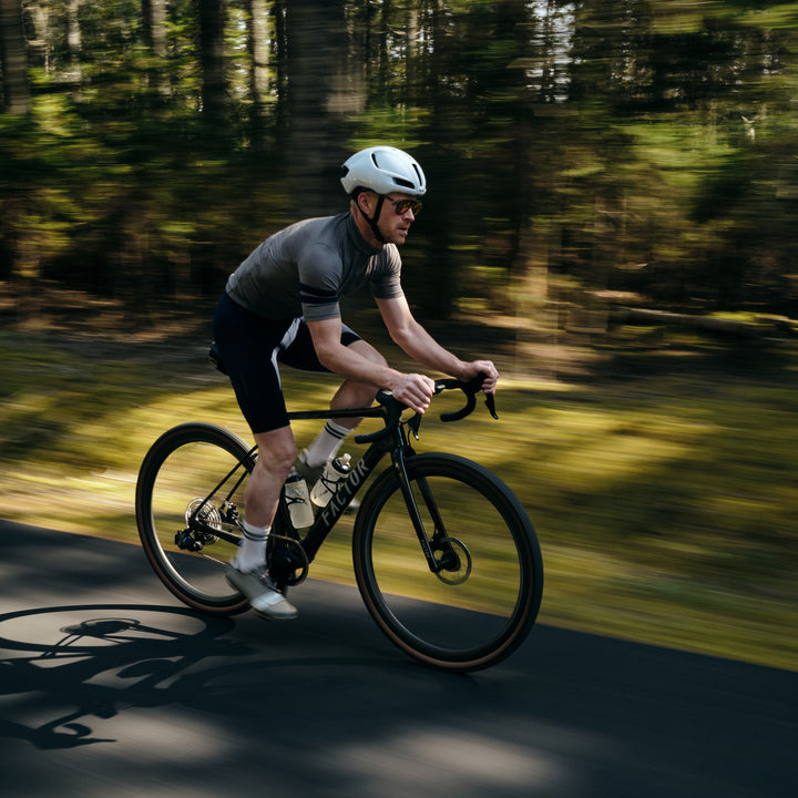 Pinebury Rangeley Long Sleeve Merino Wool Cycling Jersey in Granite. Male cyclist riding fast on a road with a pan shot of the trees in the background. He is wearing a grey jersey with a white helmet and black bib shorts, riding a Factor bike.