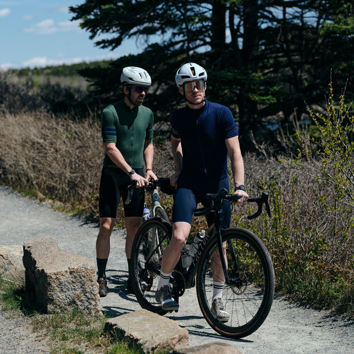 Pinebury Rangeley Long Sleeve Merino Wool Cycling Jersey in Atlantic Blue. Two male cyclists stopped on a small gravel path. Both on gravel bikes with one wearing a blue short sleeve jersey with grey strips and the other wearing a similar green jersey with blue stripes. 