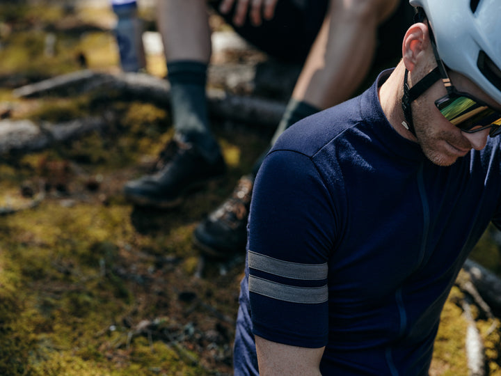 Pinebury Rangeley Short Sleeve Cycling Jersey in Atlantic Blue. Male cyclist sitting on the mossy ground close to the camera showing jersey detail with grey bicep stripes.