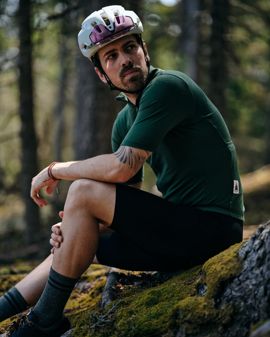 Pinebury Rangeley Long Sleeve Merino Wool Cycling Jersey in Pine. Male cyclist sitting on a mossy rock looking behind himself with his arm on his knee. He is wearing a green cycling jersey with black bibs and a white helmet.