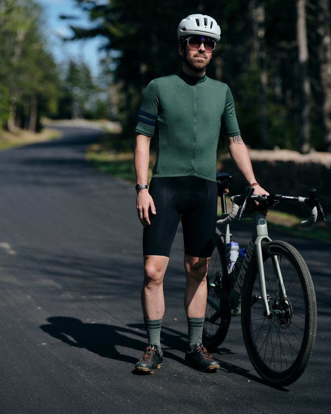 Pinebury Rangeley Long Sleeve Merino Wool Cycling Jersey in Pine. Male cyclist stopped on a road standing next to his gravel bike. Looking towards the camera wearing a green short sleeve jersey with two blue stripes on the arm, black bibs and green socks with a white helmet.