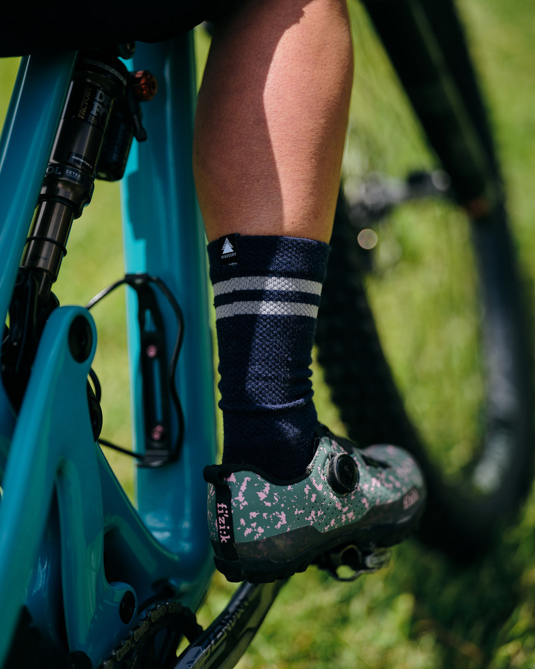 Pinebury Signature Merino Wool Sock in Navy. Close up shot of mounting biker on a teal bike, right leg on pedal with navy blue sock that has light grey stripes and they are wearing a colorful fizik shoe.