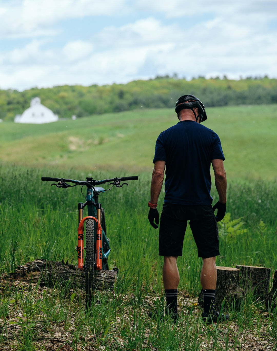 Pinebury Signature Merino Wool Sock in Navy. Male mountain biker standing with their back to the camera and bike balanced on a log facing the camera. They are standing in front of a large field wearing a navy blue tee, black shorts, and navy socks.