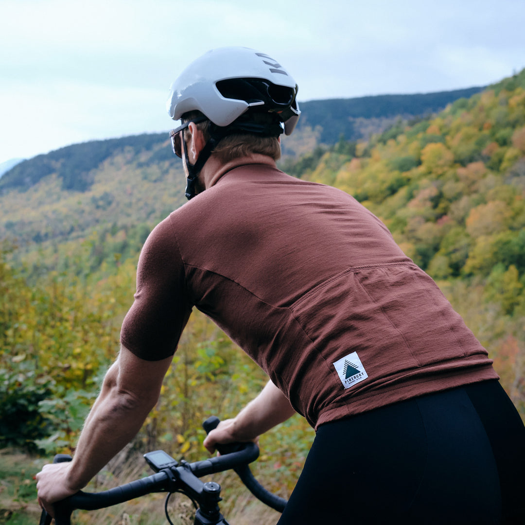 Pinebury Grafton Short Sleeve Merino Wool Cycling Jersey in Brick Red, backside view showing jersey details of man cycling in Maine with mountains