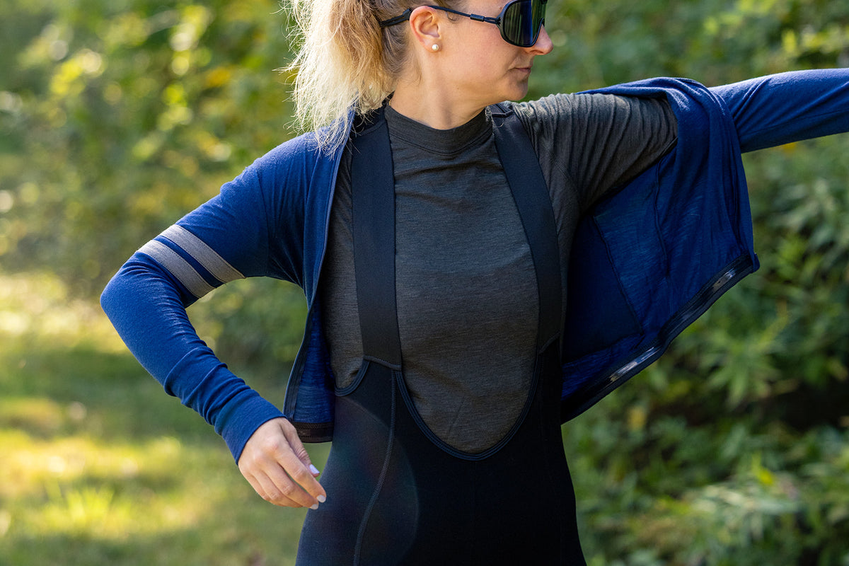A woman outdoors wearing a blue long-sleeve cycling jersey layered over a dark gray and black bib overalls, with her arm extended to show the side and back of the jersey.