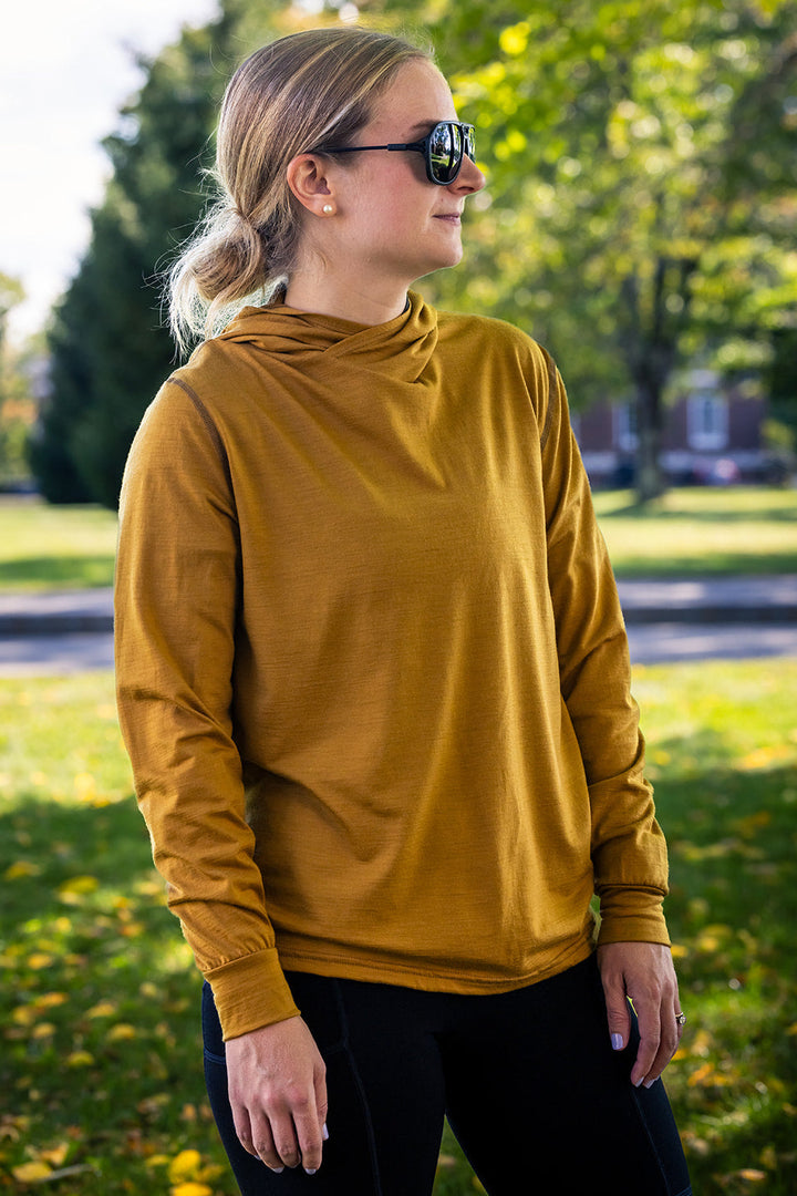 A woman wearing a mustard yellow long-sleeve high-neck top and black pants, standing outdoors in a park with trees and grass in the background.