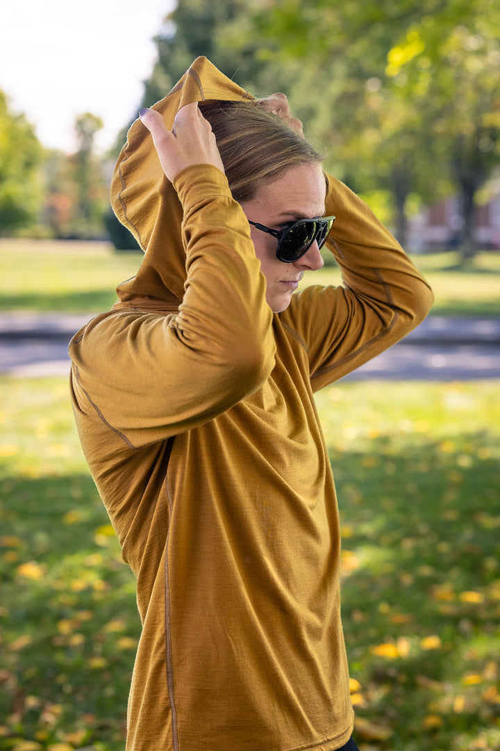 A woman wearing a mustard yellow long-sleeve athletic hoodie and black sunglasses stands in a sunny park, adjusting the hood of the sweatshirt.