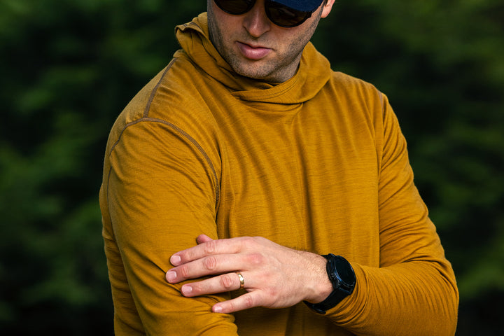 A man wearing a mustard yellow long-sleeve high-neck shirt, sunglasses, and a black watch, standing outdoors with his arms crossed.