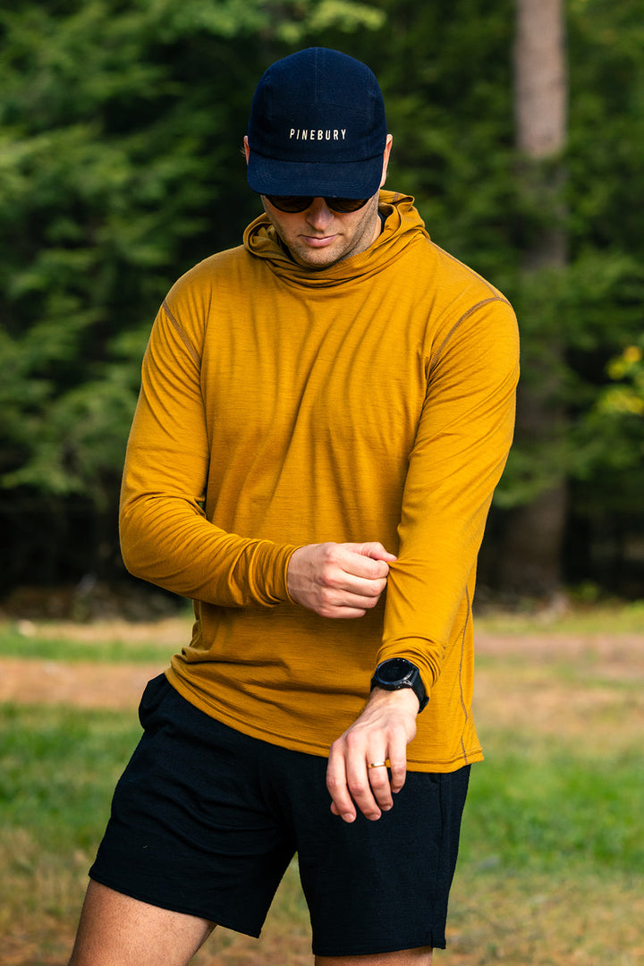 A man wearing a mustard yellow long-sleeve performance hoodie, black shorts, and a navy blue Piquadro cap stands outdoors in a grassy area, looking down at his wrist.