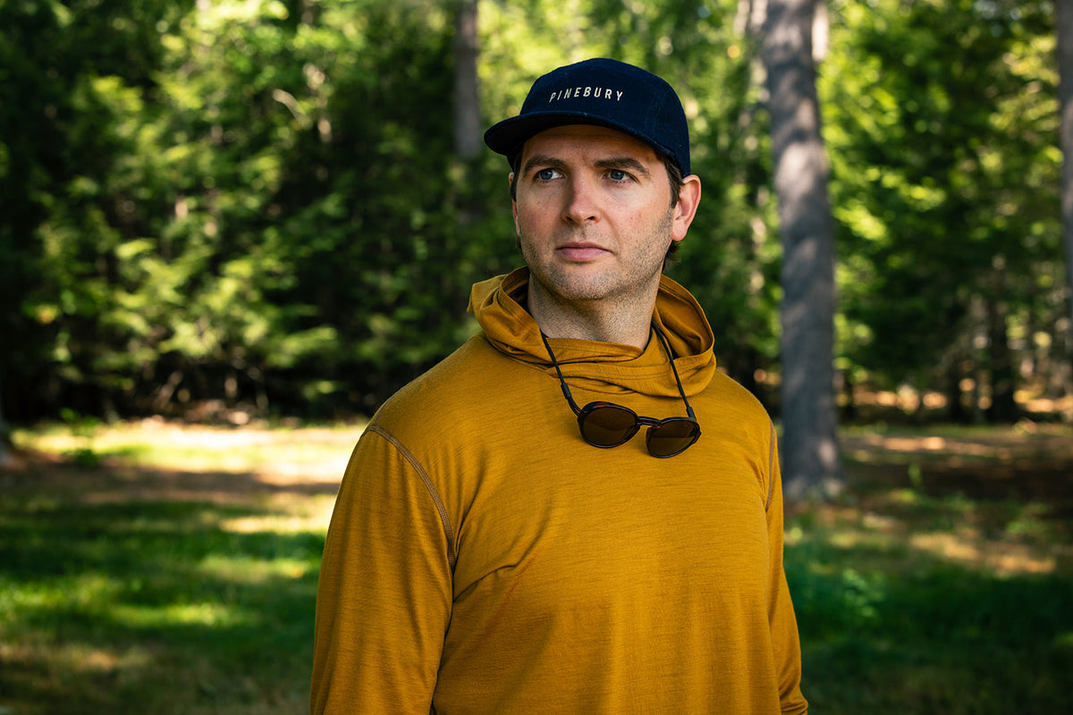 A man wearing a mustard yellow hooded sweatshirt and a dark navy cap with 'PINEBURY' embroidered on the front, standing outdoors in a sunlit forest setting.