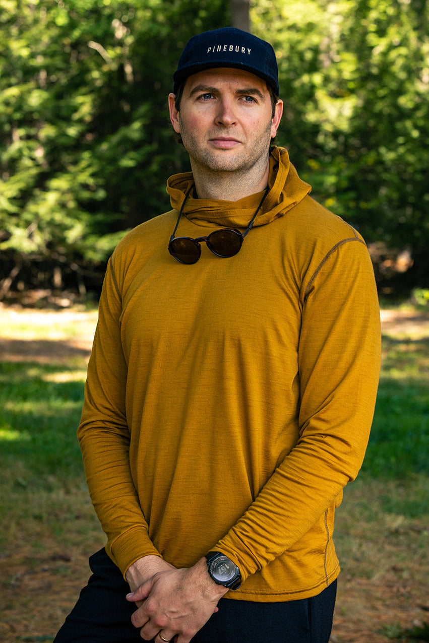 A man standing outdoors in a wooded area wearing a mustard yellow long-sleeve performance hoodie, black athletic shorts, a black cap with white text, and sunglasses hanging from the collar.