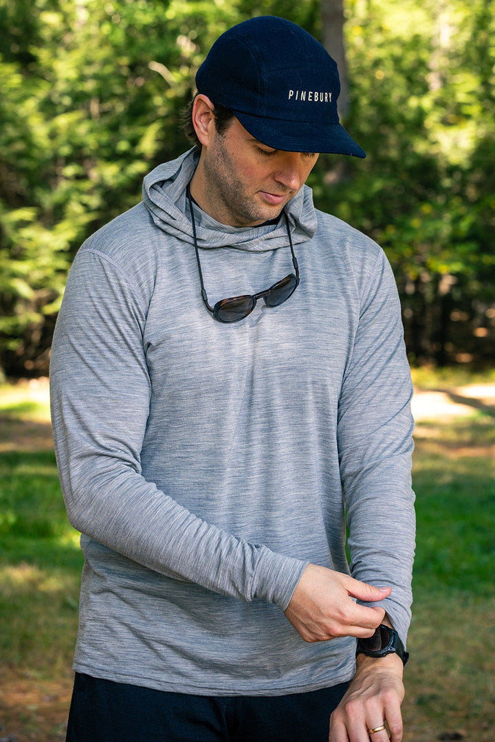 A man wearing a light gray long-sleeve performance hoodie, dark navy shorts, and a navy Piquadro cap stands outdoors in a wooded setting, looking down at his wrist.