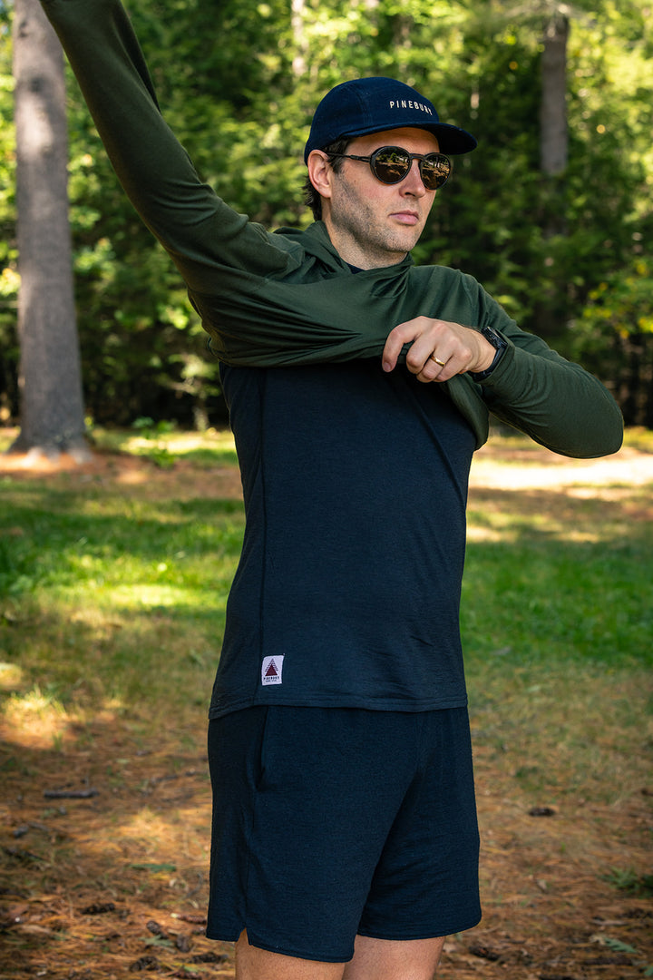 A man wearing a dark navy long-sleeve performance shirt, matching navy shorts, and a navy cap stands outdoors in a wooded setting, pulling up the sleeve of his shirt.