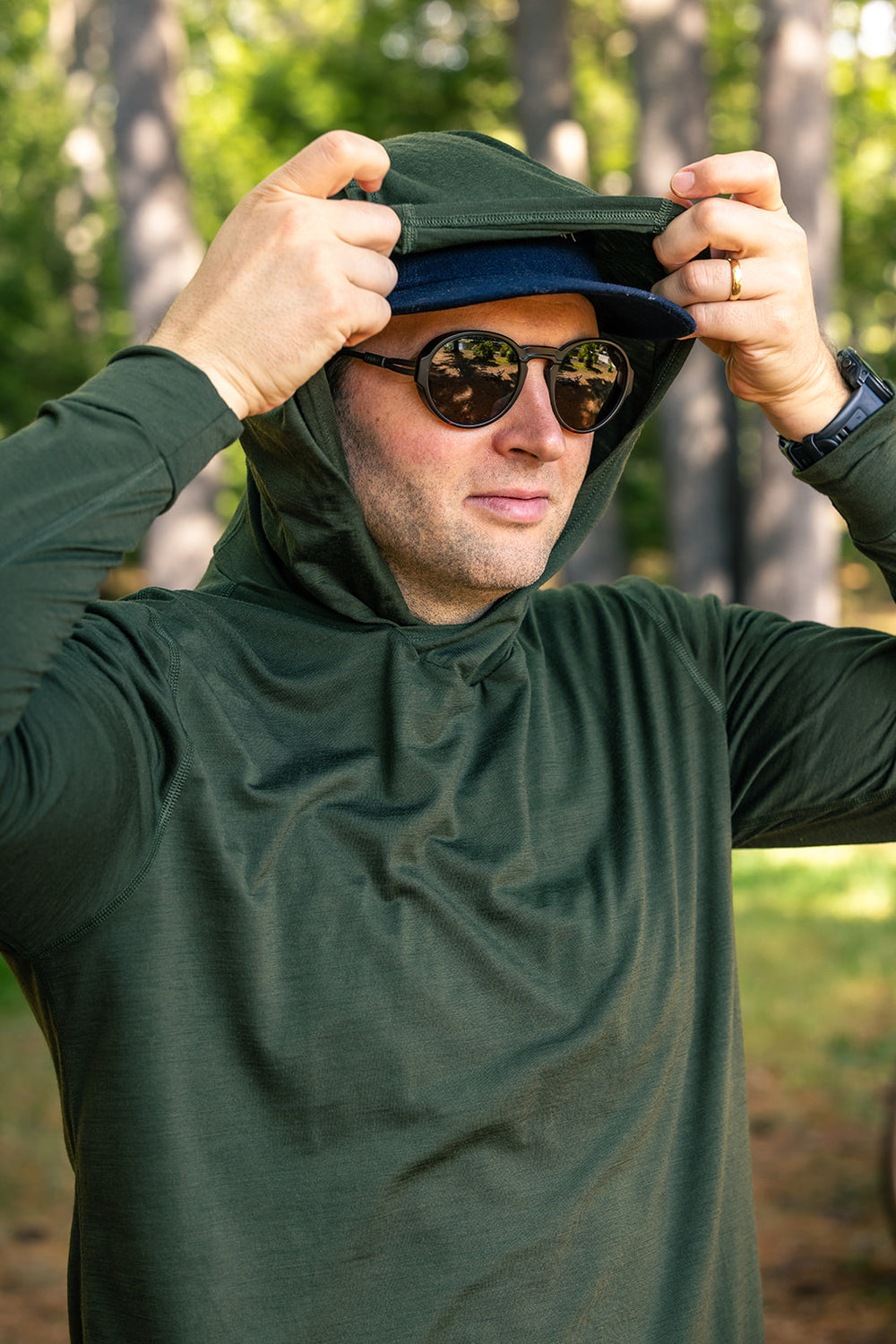 A man wearing a dark green long-sleeve hooded performance shirt and sunglasses, adjusting the hood of the shirt in an outdoor forest setting.