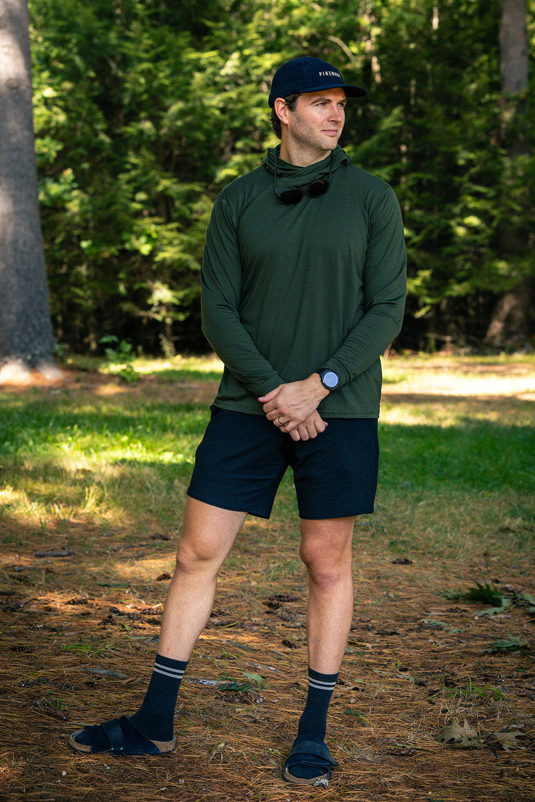 A man standing outdoors in a forest wearing an olive green long-sleeve athletic shirt, black shorts, black socks, and a black cap.