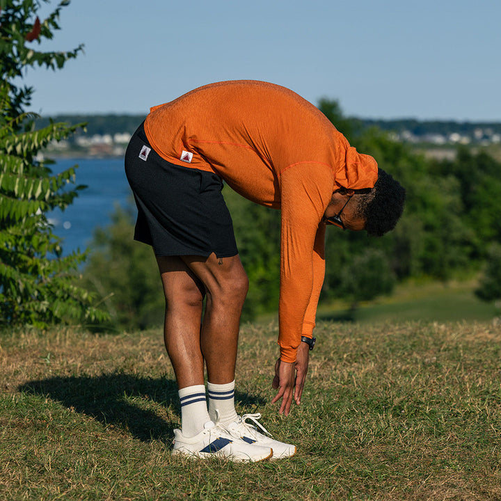 A man in an orange long-sleeve hoodie, black shorts, white socks, and white sneakers bends forward on a grassy hill with trees and water in the background.