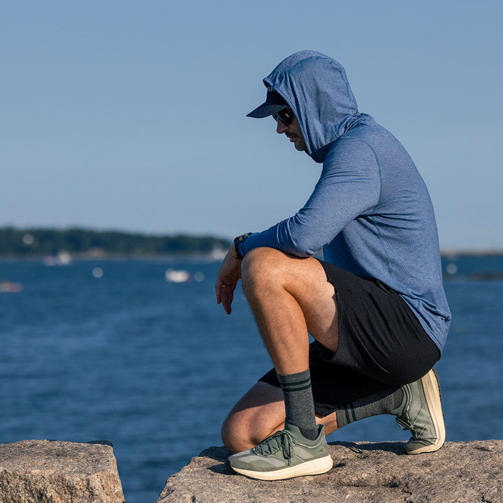 A man wearing a blue hooded long-sleeve athletic shirt, black shorts, and gray sneakers crouches on a stone pier with a body of water and distant shoreline in the background.