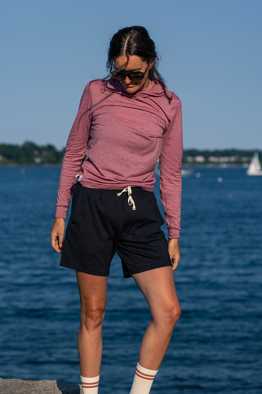 A woman wearing a dusty rose long-sleeve hooded top and black athletic shorts stands by the water with a lake and distant sailboat in the background.