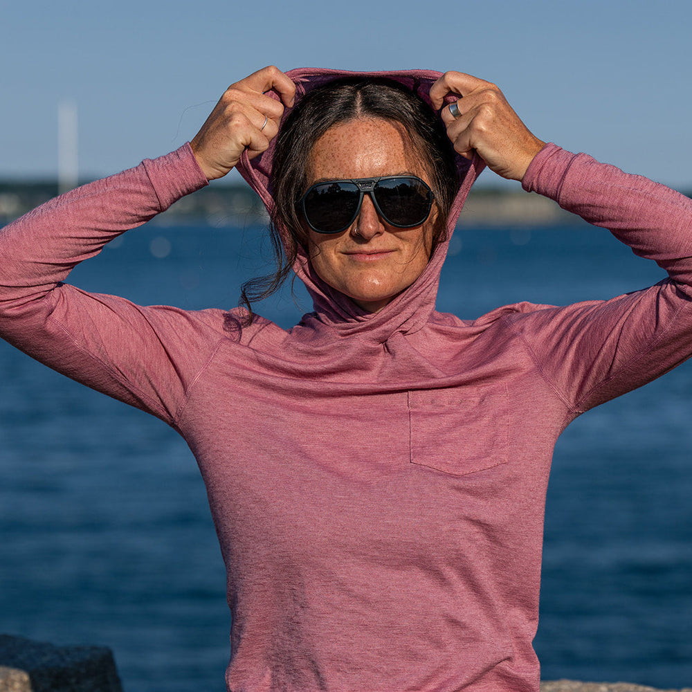 A woman wearing a dusty pink long-sleeve hooded top and black sunglasses stands outdoors by the water, adjusting the hood of her shirt.