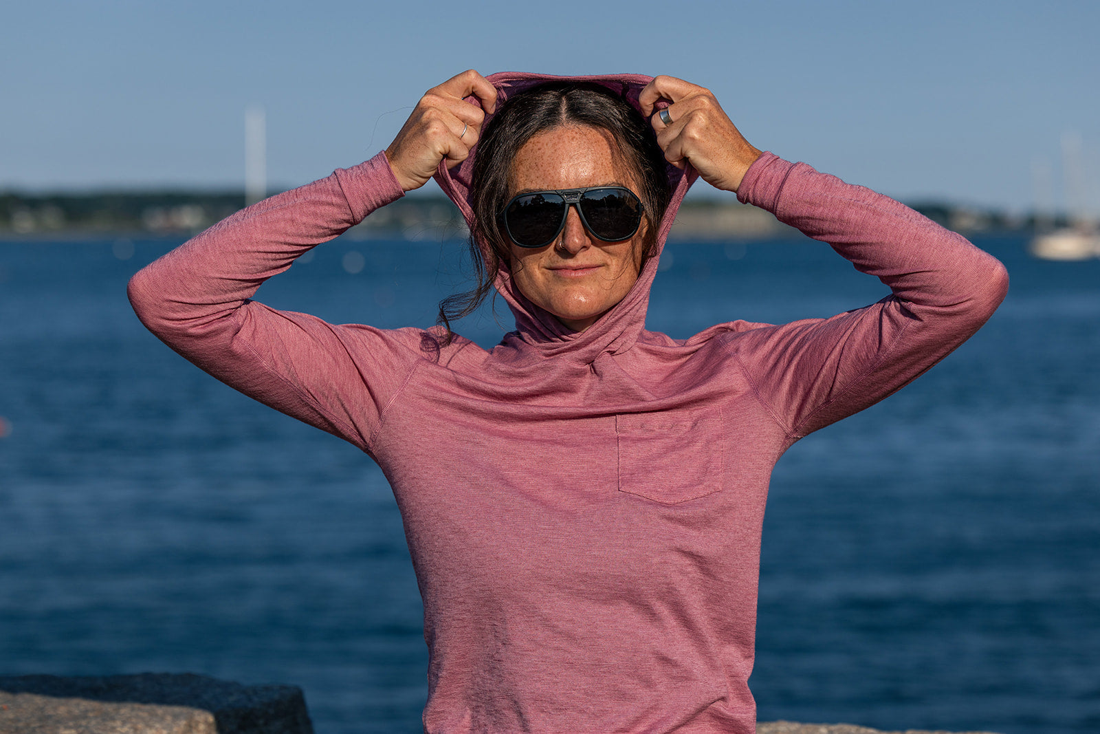 A woman wearing a dusty pink long-sleeve hooded top and black sunglasses stands outdoors by the water, adjusting the hood of her shirt.
