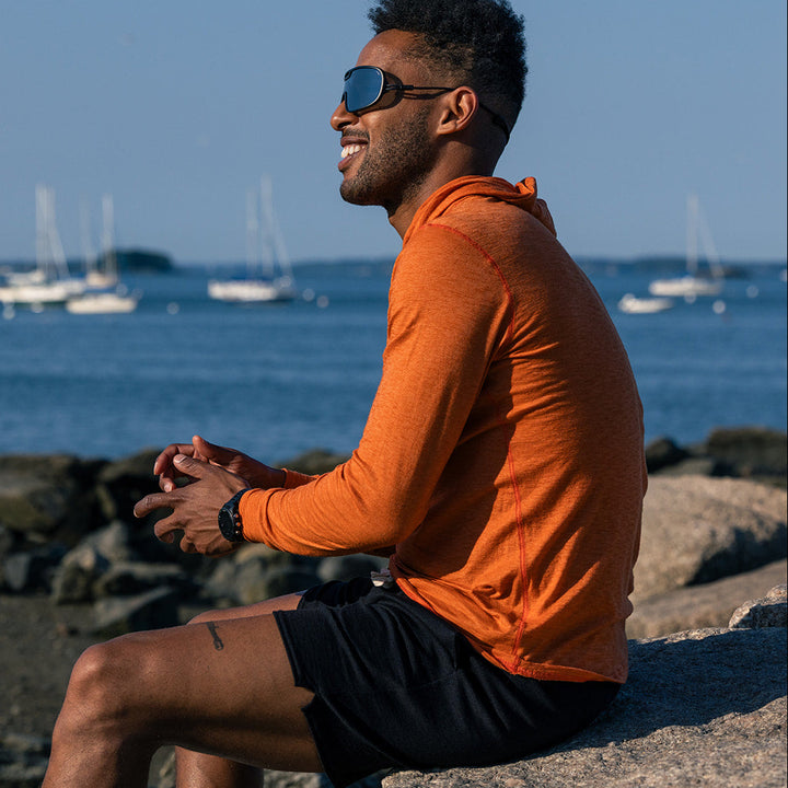 A man wearing an orange long-sleeve athletic shirt and black shorts sits on a rocky shoreline, looking out toward the water with sailboats in the distance.