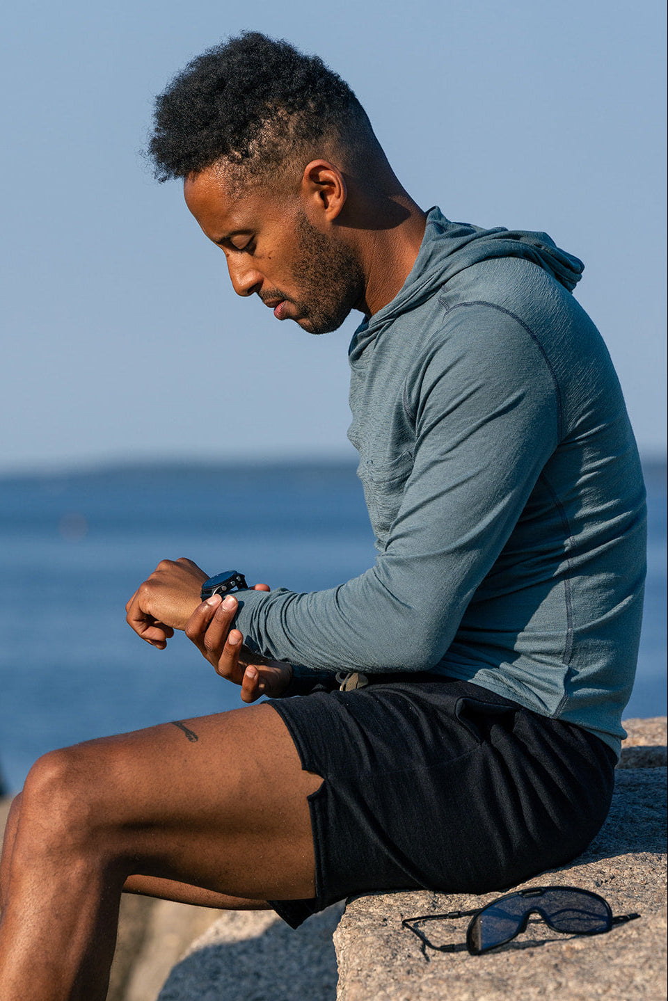 A man in a slate blue long-sleeve athletic hoodie and black shorts sits on a rock by the ocean, checking his watch. A pair of sunglasses rests on the rock beside him.