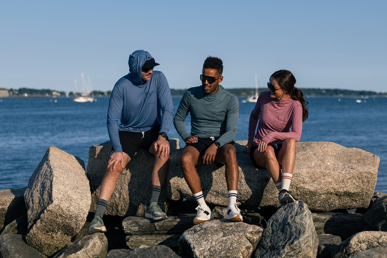 Three people sitting on large rocks by the water wearing matching athletic long-sleeve tops in blue, gray, and pink, with a harbor and boats in the background.