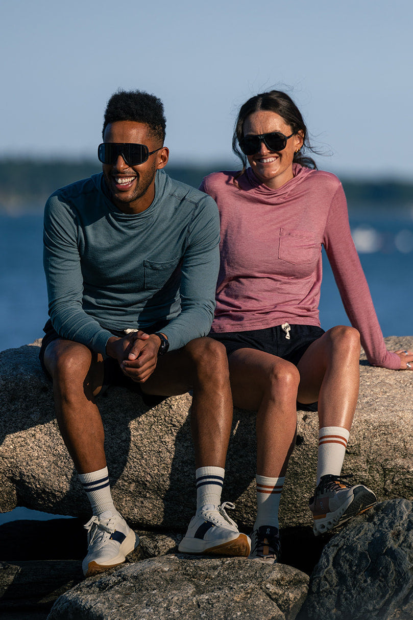 A man and a woman sitting on large rocks outdoors by the water, wearing sunglasses, athletic long-sleeve tops, shorts, socks, and sneakers.