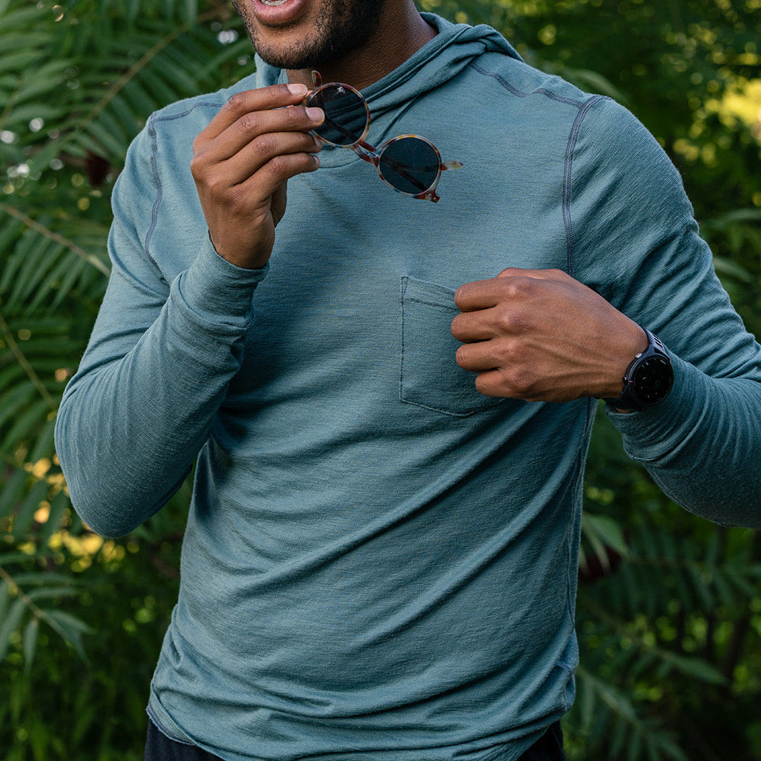 A man wearing a teal blue long-sleeve performance hoodie with a chest pocket, holding a pair of sunglasses in front of his face, set against a lush green outdoor background.