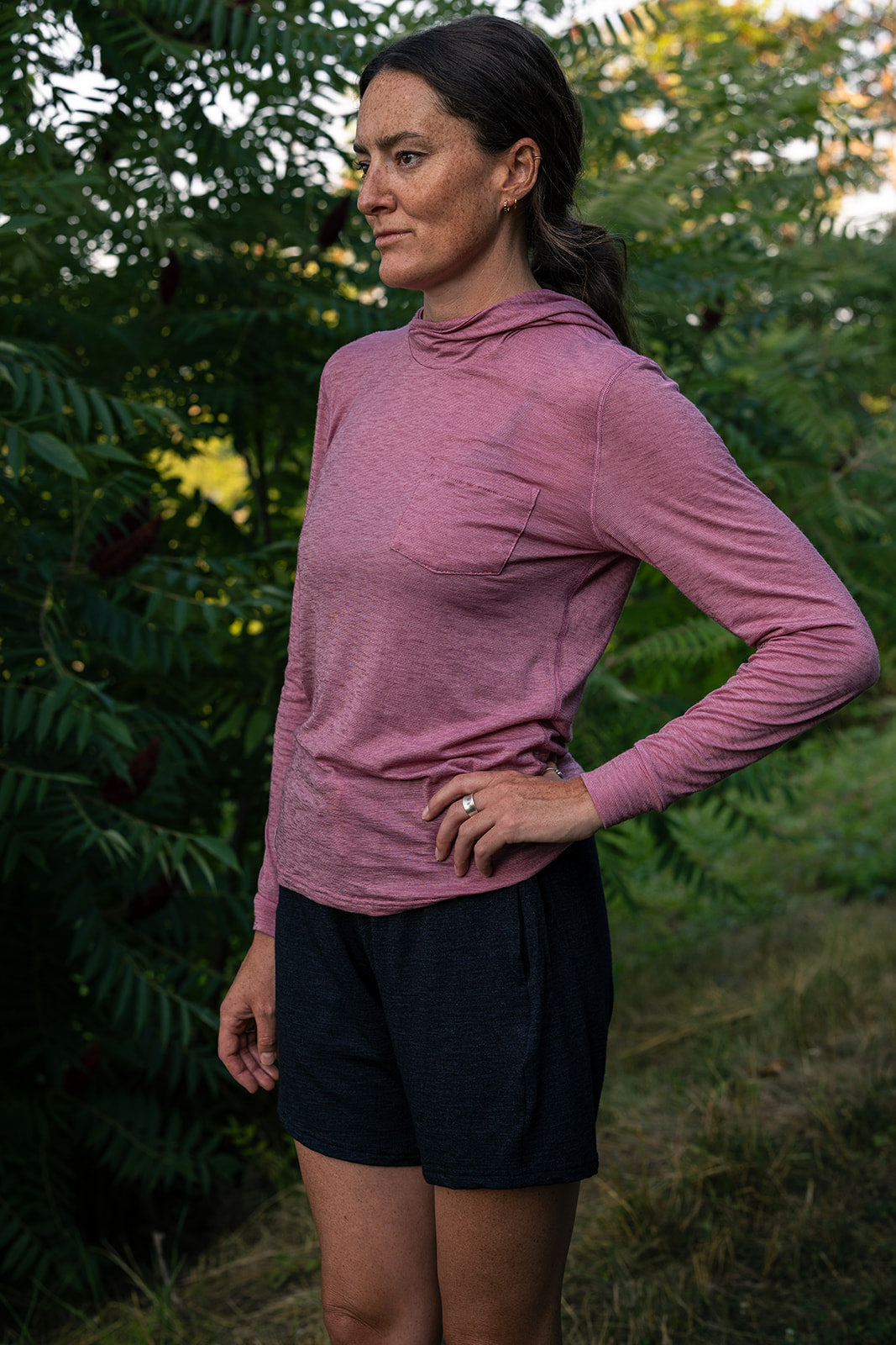 A woman wearing a dusty rose long-sleeve athletic hoodie and dark charcoal shorts, standing outdoors in front of green foliage.